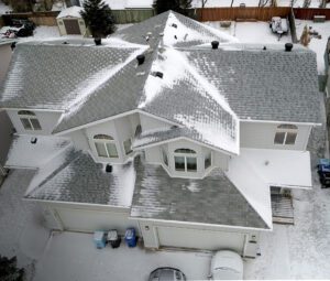 Aerial view of a snow-covered house with a partially cleared gray shingle roof.